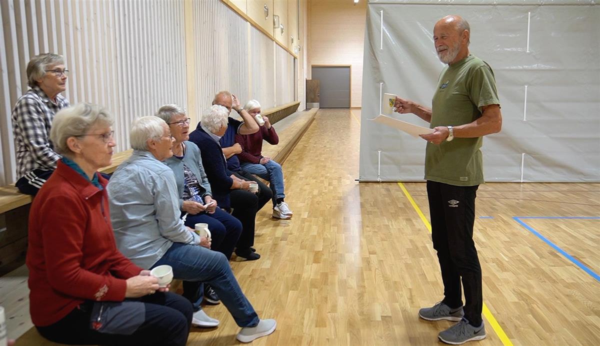 Mann står foran andre seniorer med et ark i hånda og prater - Klikk for stort bilde